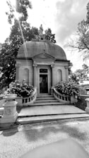 A historic mausoleum with a domed roof stands prominently amidst a backdrop of trees. The structure features grand stone steps leading up to an ornate door, flanked by large flowering bushes. A statue is perched atop the dome, and intricate stone carvings adorn the exterior walls.