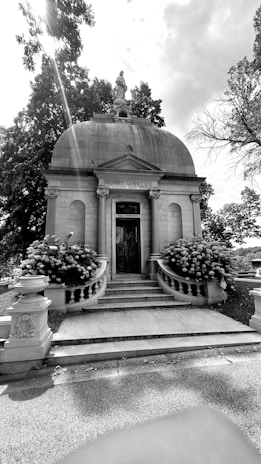 A historic mausoleum with a domed roof stands prominently amidst a backdrop of trees. The structure features grand stone steps leading up to an ornate door, flanked by large flowering bushes. A statue is perched atop the dome, and intricate stone carvings adorn the exterior walls.
