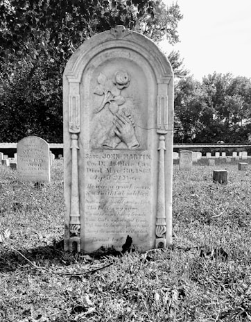 A historic cemetery with an old tombstone prominently placed in the foreground, intricately carved with symbols and inscriptions. In the background, other tombstones populate the green grass, partially shaded by tall trees, creating a peaceful and solemn atmosphere.