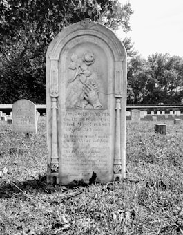 A historic cemetery with an old tombstone prominently placed in the foreground, intricately carved with symbols and inscriptions. In the background, other tombstones populate the green grass, partially shaded by tall trees, creating a peaceful and solemn atmosphere.