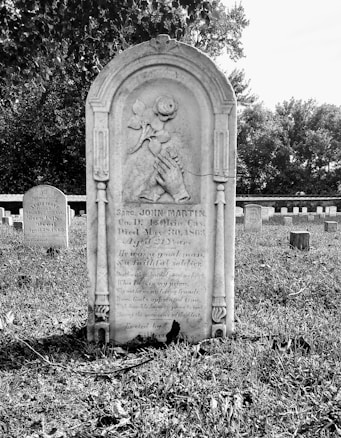 A historic cemetery with an old tombstone prominently placed in the foreground, intricately carved with symbols and inscriptions. In the background, other tombstones populate the green grass, partially shaded by tall trees, creating a peaceful and solemn atmosphere.