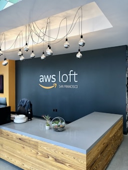 A modern reception area featuring a sleek wooden desk with a light gray countertop. Hanging above are several decorative light bulbs with black cords creating an artistic display. The wall behind the desk is dark gray with the logo 'aws loft San Francisco' in white and orange branding. The desk is adorned with a glass terrarium and a small plant, adding a touch of nature to the contemporary setting.