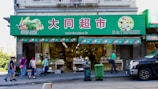A busy street market scene with people walking in front of a grocery store. The storefront is adorned with a green sign featuring images of vegetables and fruits, and shelves displaying various produce. Pedestrians are seen carrying bags and pushing carts along the sidewalk.