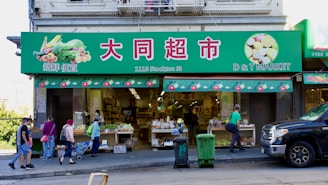 A busy street market scene with people walking in front of a grocery store. The storefront is adorned with a green sign featuring images of vegetables and fruits, and shelves displaying various produce. Pedestrians are seen carrying bags and pushing carts along the sidewalk.