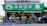 A busy street market scene with people walking in front of a grocery store. The storefront is adorned with a green sign featuring images of vegetables and fruits, and shelves displaying various produce. Pedestrians are seen carrying bags and pushing carts along the sidewalk.