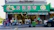 A busy street market scene with people walking in front of a grocery store. The storefront is adorned with a green sign featuring images of vegetables and fruits, and shelves displaying various produce. Pedestrians are seen carrying bags and pushing carts along the sidewalk.