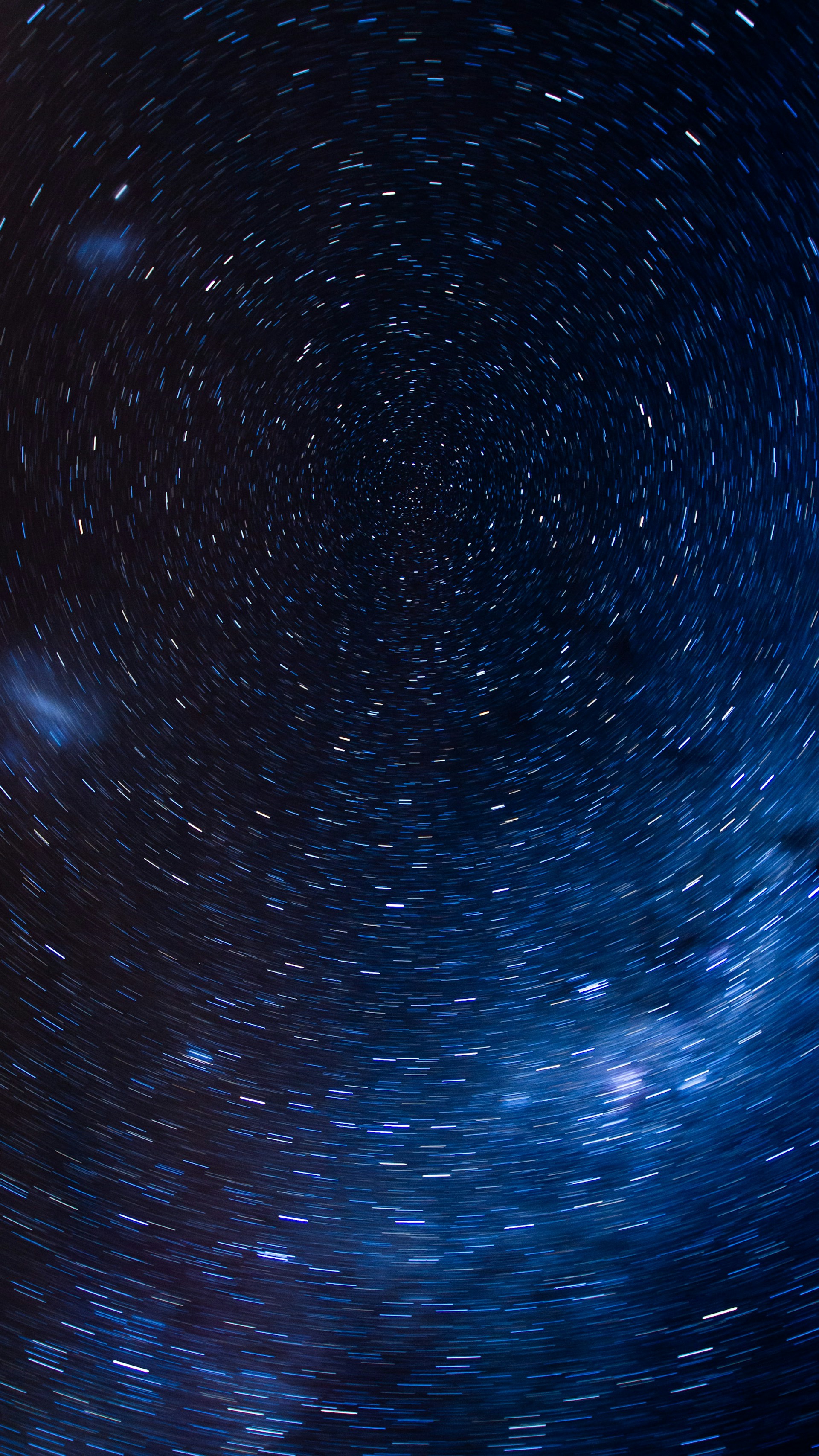A mesmerizing view of the night sky with stars creating a swirling effect, suggesting a long-exposure photograph capturing the Earth's rotation. The stars are predominantly white and blue in color against a deep blue-black backdrop, giving a sense of motion and depth.