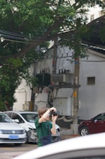 A person in a green shirt is using a camera to take photos in an outdoor urban setting. Nearby, there are several cars parked, including a white car with a minor front-end collision. The background includes an electrical installation with wires and equipment, and a large tree providing some greenery.