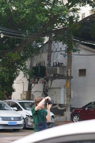 A person in a green shirt is using a camera to take photos in an outdoor urban setting. Nearby, there are several cars parked, including a white car with a minor front-end collision. The background includes an electrical installation with wires and equipment, and a large tree providing some greenery.