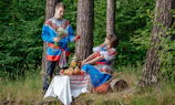 A couple dressed in traditional colorful clothing is in a forest setting. The man is holding a bouquet and an apple, while the woman sits playfully on a log next to a small table covered with a patterned cloth. The table holds traditional items, including a samovar and various breads.