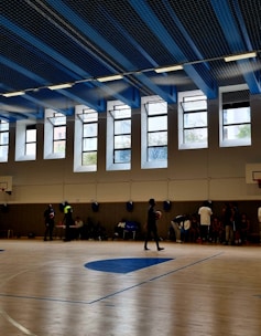 A large indoor basketball court with clean wooden flooring and multiple people present. The ceiling is high with blue and black panels and fluorescent lights. Tall windows allow natural light to fill the space. Several basketball hoops are visible, and individuals appear to be engaged in different activities, with some holding basketballs.