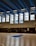 A large indoor basketball court with clean wooden flooring and multiple people present. The ceiling is high with blue and black panels and fluorescent lights. Tall windows allow natural light to fill the space. Several basketball hoops are visible, and individuals appear to be engaged in different activities, with some holding basketballs.
