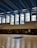 A large indoor basketball court with clean wooden flooring and multiple people present. The ceiling is high with blue and black panels and fluorescent lights. Tall windows allow natural light to fill the space. Several basketball hoops are visible, and individuals appear to be engaged in different activities, with some holding basketballs.
