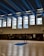 A large indoor basketball court with clean wooden flooring and multiple people present. The ceiling is high with blue and black panels and fluorescent lights. Tall windows allow natural light to fill the space. Several basketball hoops are visible, and individuals appear to be engaged in different activities, with some holding basketballs.