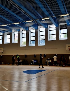 A large indoor basketball court with clean wooden flooring and multiple people present. The ceiling is high with blue and black panels and fluorescent lights. Tall windows allow natural light to fill the space. Several basketball hoops are visible, and individuals appear to be engaged in different activities, with some holding basketballs.