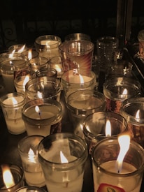 A collection of lit candles in glass holders creates a warm, flickering light against a dark background. Some candles have labels or images printed on the glass.