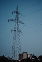 A tall lattice power transmission tower stands prominently against a backdrop of a clear evening sky. The structure is framed with multiple intersecting metal beams and wires extending outward. Below the tower, there is a partially visible block of residential buildings and silhouettes of trees, suggesting an urban environment.