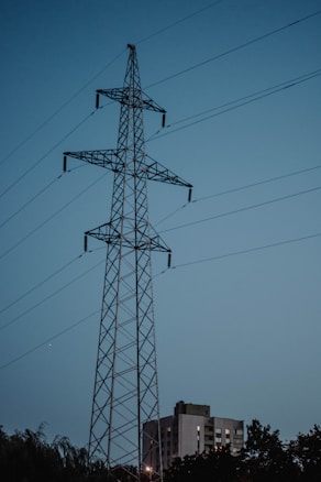 A tall lattice power transmission tower stands prominently against a backdrop of a clear evening sky. The structure is framed with multiple intersecting metal beams and wires extending outward. Below the tower, there is a partially visible block of residential buildings and silhouettes of trees, suggesting an urban environment.