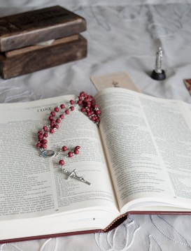 An open book, likely a Bible, is centered on a decorative fabric surface. A rosary with red beads and a silver crucifix is placed on top of the pages. To the left, there are stacked wooden boxes, and in the background, there are blurred religious figurines and cards.