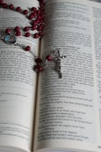 An open book with text on its pages, displaying part of a religious scripture. A rosary with red beads and a silver crucifix is laid across the pages. A small medallion with an image is attached to the rosary.