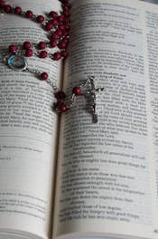 An open book with text on its pages, displaying part of a religious scripture. A rosary with red beads and a silver crucifix is laid across the pages. A small medallion with an image is attached to the rosary.