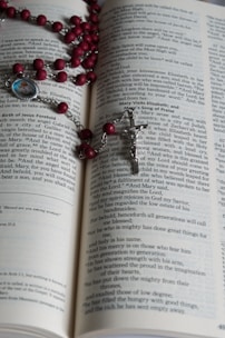 An open book with text on its pages, displaying part of a religious scripture. A rosary with red beads and a silver crucifix is laid across the pages. A small medallion with an image is attached to the rosary.