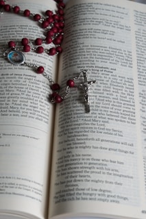 An open book with text on its pages, displaying part of a religious scripture. A rosary with red beads and a silver crucifix is laid across the pages. A small medallion with an image is attached to the rosary.
