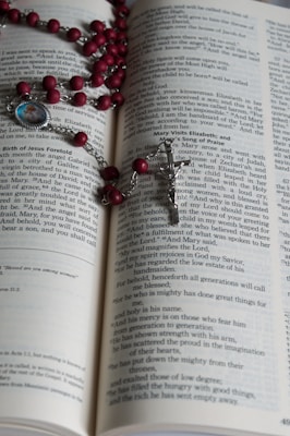 An open book with text on its pages, displaying part of a religious scripture. A rosary with red beads and a silver crucifix is laid across the pages. A small medallion with an image is attached to the rosary.