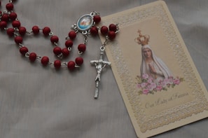 A rosary with red beads and a silver crucifix lies next to a prayer card featuring an image of Our Lady of Fatima, adorned with a crown and surrounded by pink roses. The card has intricate lace-like borders and soft colors.