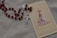 A rosary with red beads and a silver crucifix lies next to a prayer card featuring an image of Our Lady of Fatima, adorned with a crown and surrounded by pink roses. The card has intricate lace-like borders and soft colors.