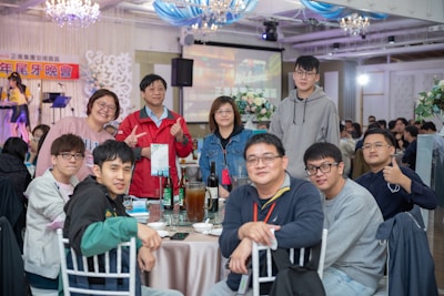 A group of eleven people are gathered around a table in a well-lit indoor venue. Some are standing behind the table while others are seated. The background features decorations, including chandeliers, and there is a stage with musical equipment and a backdrop with text. The table is set with plates, glasses, bottles, and pitchers.