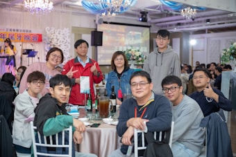 A group of eleven people are gathered around a table in a well-lit indoor venue. Some are standing behind the table while others are seated. The background features decorations, including chandeliers, and there is a stage with musical equipment and a backdrop with text. The table is set with plates, glasses, bottles, and pitchers.