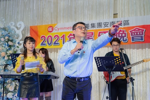A group of people is on stage at an event. The central figure is a man in a blue shirt holding a microphone, gesturing with his hand. A woman wearing a yellow top and black skirt stands to his side, also holding a microphone and papers. In the background, a man with a guitar stands by a music stand, along with a person behind a keyboard. The backdrop features a banner with Chinese characters and the year '2021'. The setting is decorated with flowers and lights, suggesting a formal or celebratory occasion.