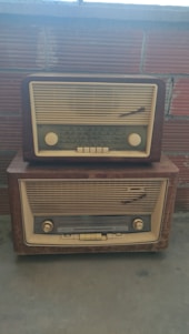 Two vintage radios with cream and brown tones are stacked on top of each other against a brick wall backdrop. The radios feature dials and frequency indicators typical of mid-20th century designs.