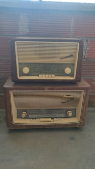 Two vintage radios with cream and brown tones are stacked on top of each other against a brick wall backdrop. The radios feature dials and frequency indicators typical of mid-20th century designs.