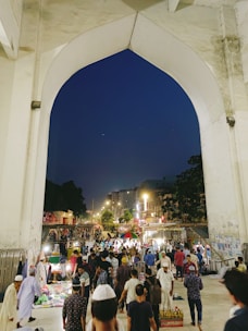 A bustling market scene under a large stone archway with numerous people walking and interacting. Stalls are set up with various items for sale, and a variety of vendors and shoppers can be seen. The background shows a city street lined with lights and buildings, suggesting an urban setting during nighttime.