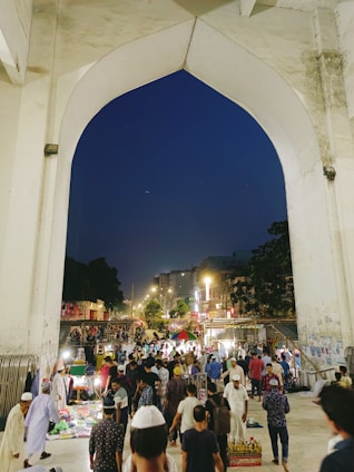 A bustling market scene under a large stone archway with numerous people walking and interacting. Stalls are set up with various items for sale, and a variety of vendors and shoppers can be seen. The background shows a city street lined with lights and buildings, suggesting an urban setting during nighttime.