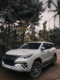 a white car parked on a dirt road with trees and a building in the background