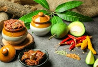 A rustic arrangement of culinary ingredients features ceramic pots with spicy pickles, fresh mangoes, a bunch of red chili peppers, turmeric, and various seeds on a gray stone surface. The background includes green leaves, pieces of wood, and burlap fabric, creating a natural and earthy setting.