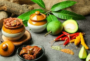 A rustic arrangement of culinary ingredients features ceramic pots with spicy pickles, fresh mangoes, a bunch of red chili peppers, turmeric, and various seeds on a gray stone surface. The background includes green leaves, pieces of wood, and burlap fabric, creating a natural and earthy setting.