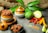 A rustic arrangement of culinary ingredients features ceramic pots with spicy pickles, fresh mangoes, a bunch of red chili peppers, turmeric, and various seeds on a gray stone surface. The background includes green leaves, pieces of wood, and burlap fabric, creating a natural and earthy setting.