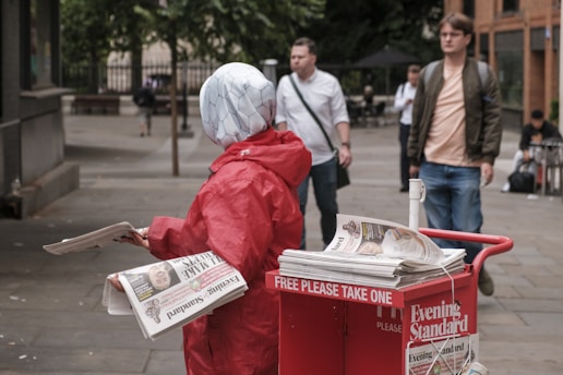 A person in a red jacket is handing out newspapers from a red cart labeled 'Evening Standard' on a street. The person's head is covered with a white patterned headscarf. In the background, there are several pedestrians and buildings.