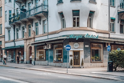 A corner building with a storefront featuring signs for 'Marja Entrich' and 'stureplanskliniken'. The facade is white with green accents, and the area includes large windows. Two people are riding scooters on the sidewalk, and a few pedestrians are walking nearby.