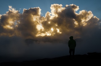 A dramatic scene with a silhouette of a person standing against a backdrop of thick, illuminated clouds. The light breaks through the clouds, casting a golden glow amidst the dark atmosphere. Mist surrounds the figure, enhancing the mysterious and contemplative mood.