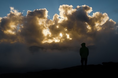 A dramatic scene with a silhouette of a person standing against a backdrop of thick, illuminated clouds. The light breaks through the clouds, casting a golden glow amidst the dark atmosphere. Mist surrounds the figure, enhancing the mysterious and contemplative mood.