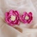 Two handcrafted pink flowers with intricate petal designs are placed on a soft beige fabric background. The fabric has a delicate texture with small pearls scattered across its surface.