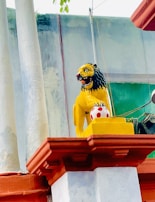 A vibrantly painted lion statue is positioned on a decorative plinth. The lion is yellow with a fierce expression, black mane, and red mouth, sitting above a red and white spotted ball. The statue is situated against a building with colorful pillars and a green section in the background.