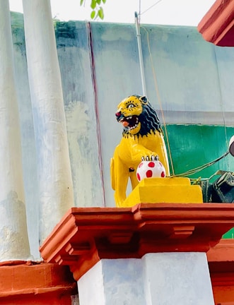 A vibrantly painted lion statue is positioned on a decorative plinth. The lion is yellow with a fierce expression, black mane, and red mouth, sitting above a red and white spotted ball. The statue is situated against a building with colorful pillars and a green section in the background.