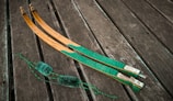 Two curved wooden objects with green and brown hues rest on a wooden plank surface. The items appear to be archery bow limbs with intricate patterns. A coiled green string with fluffy sections is positioned nearby.