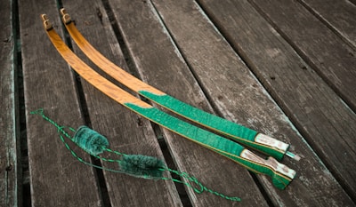Two curved wooden objects with green and brown hues rest on a wooden plank surface. The items appear to be archery bow limbs with intricate patterns. A coiled green string with fluffy sections is positioned nearby.